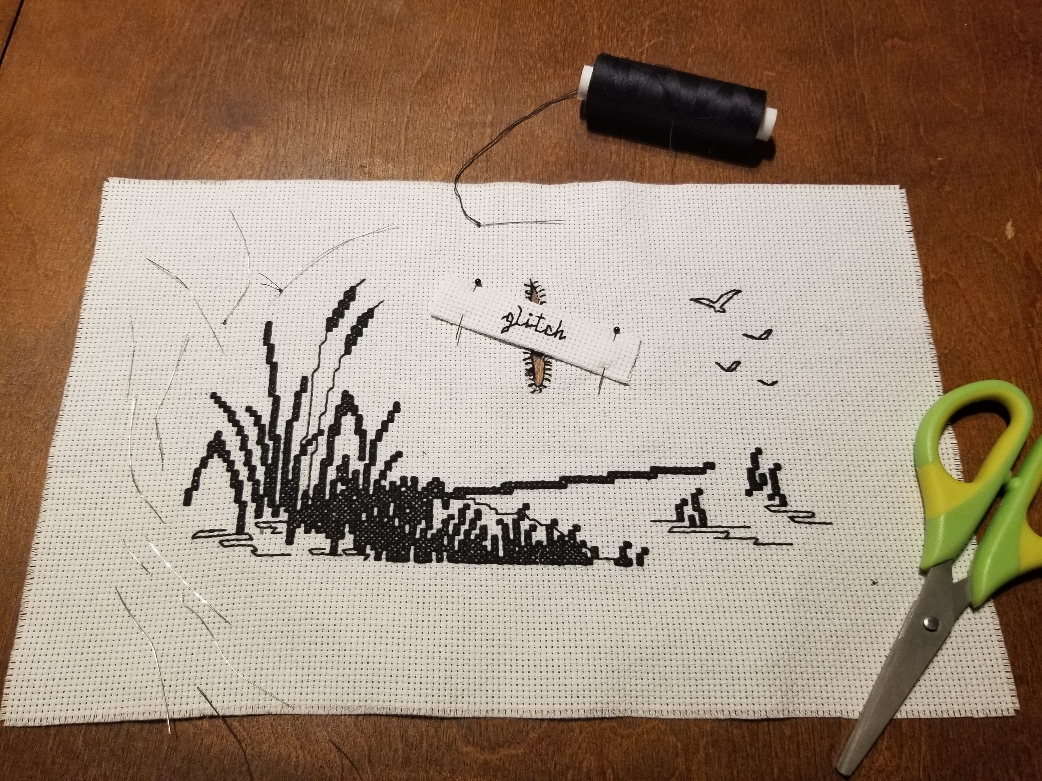 jenna moran's stitch A cross stitch that depicts cattails, surrounded by broken and bent sewing needles and a pair of scissors. A large tear in the center of the fabric has had a smaller piece of fabric that says "glitch" pinned over it. The final sewing needle has a length of thread attaching it to a spool full of thread yet to be used.
